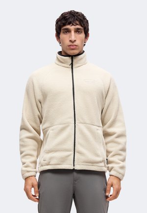 Homme portant une veste polaire beige zippée avec un col montant et un pantalon gris, debout devant un fond clair uni.