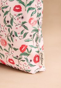 Rózsaszín szövettasak virág- és ajakmintákkal, zöld levelekkel, piros ajkakkal és rózsaszín virágokkal. Oldalán márkázott címkével.