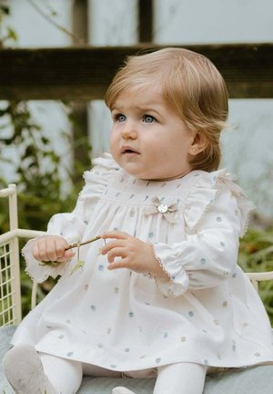 Weißes Kleid mit bunten Punkten, Rüschensaum und Kurzärmeln. Kind sitzt und hält einen Zweig. Sanfter Hintergrund mit Grün. 