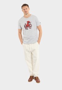 T-shirt gris à manches courtes avec un grand graphique de pieuvre rouge et noire, associé à un pantalon crème et des chaussures à lacets brunes.