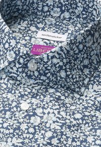 Chemise à motif floral bleu avec boutons blancs et col, portant une étiquette de marque "seidensticker" et une étiquette de tissu violette "Liberty" à l'intérieur.