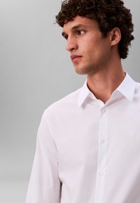 Valge nööbitav särk, mis on valmistatud siidisest kangast, terava kraega ja peene rinnal olevaga logoga; omab sirget lõiget ja klassikalist disaini.