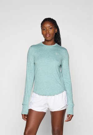 Lys teal langermet treningsskjorte med rund hals og tommelhull, kombinert med hvite shorts. Stoffet virker glatt med en svak tekstur.