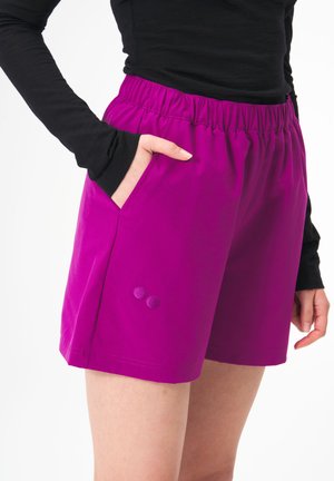 Lila Sportshorts mit elastischem Bund, seitlichen Taschen und zwei dekorativen lila Knöpfen auf der unteren rechten Seite.