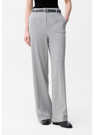 Osoba ubrana w jasnoszare spodnie o szerokich nogawkach z paskiem i czarne mokasyny, stojąca z rękami w kieszeniach na białym tle.