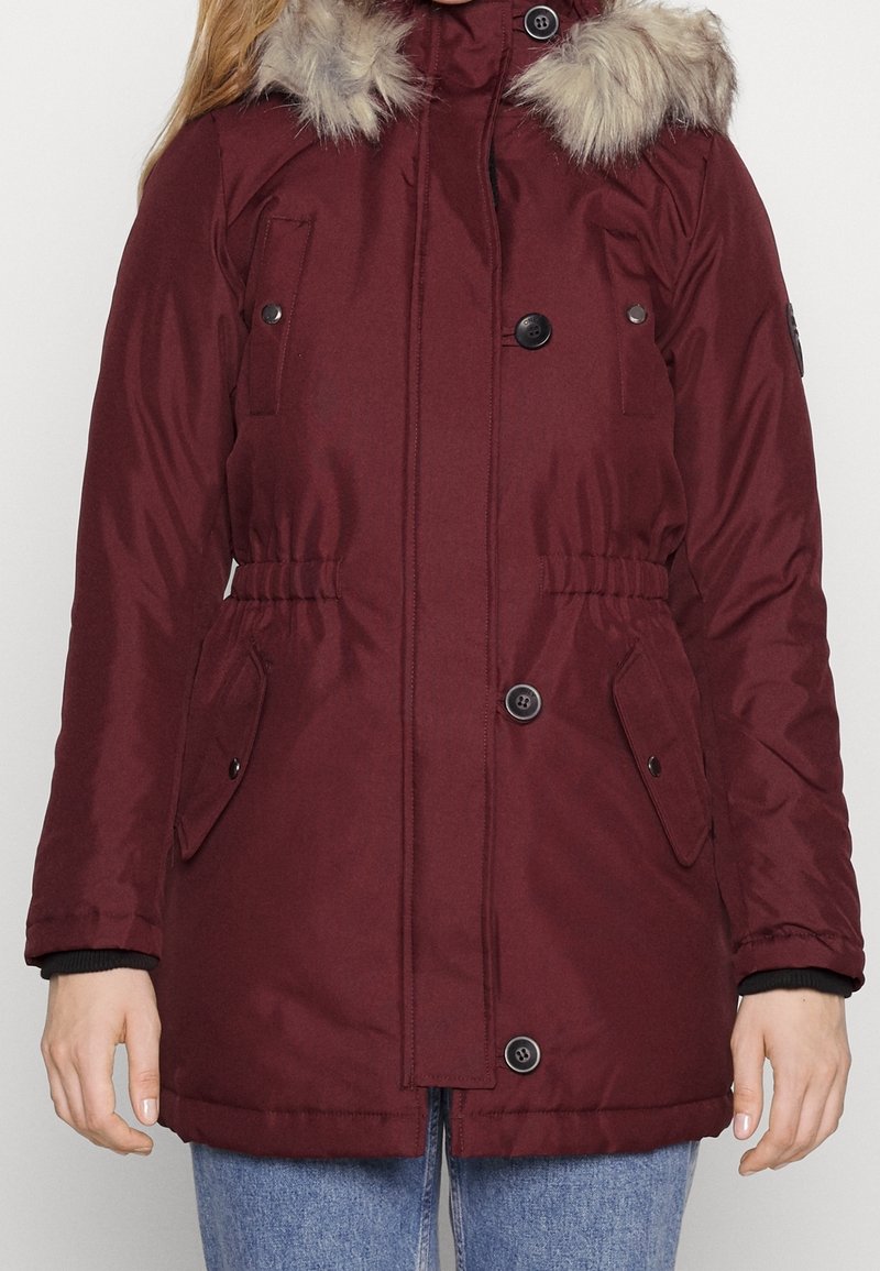 Parka bordeaux avec un col en fausse fourrure, des boutons, une taille élastique et deux poches latérales, présentant un tissu lisse et texturé.
