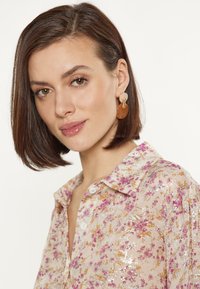 Lichtroze bloezenblouse met bloemenpatroon en metallic accenten, voorzien van een kraag en knopen aan de voorkant. Het model draagt oranje harsdruppel oorbellen.