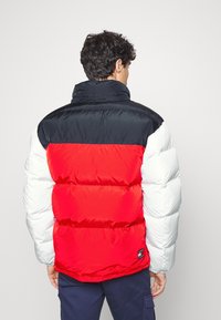 Chaqueta tipo puff con un diseño en bloque de color que presenta secciones en negro, rojo y blanco. Incluye un cuello alto y puños elásticos. Textura suave y brillante.