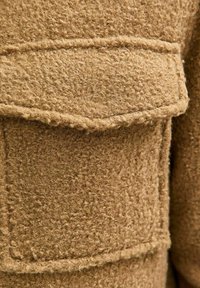 Bruine textuurgeweven wol stof met een fuzzy oppervlak, voorzien van een grote patch zak en stiksels. Het materiaal lijkt zacht en warm.