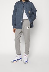 Marinfärgad vadderad jacka med tryckknappar, grå sweatpants med en vit Jordan-logotyp, och vita sneakers med blå och orange accenter.