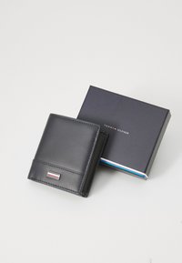 Svart lærwallet med jevn tekstur, som har sølvfargede detaljer og en liten stripe. Leveres sammen med en blå boks med logo.