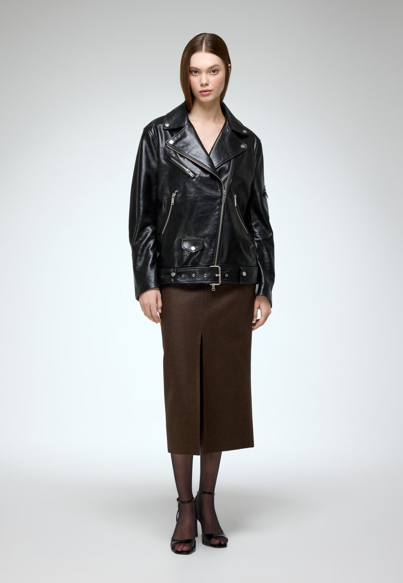 Black leather biker jacket with silver zippers, broad collar, and side pockets, paired with a brown knee-length skirt and black strappy heels.