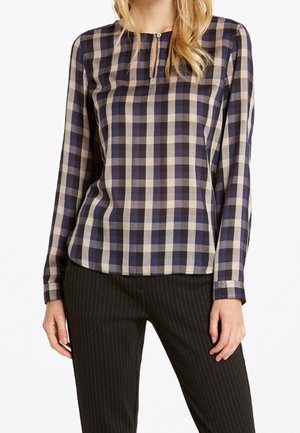 Femme portant un chemisier à manches longues à carreaux violet et beige avec un petit bouton devant et un pantalon noir à fines rayures.