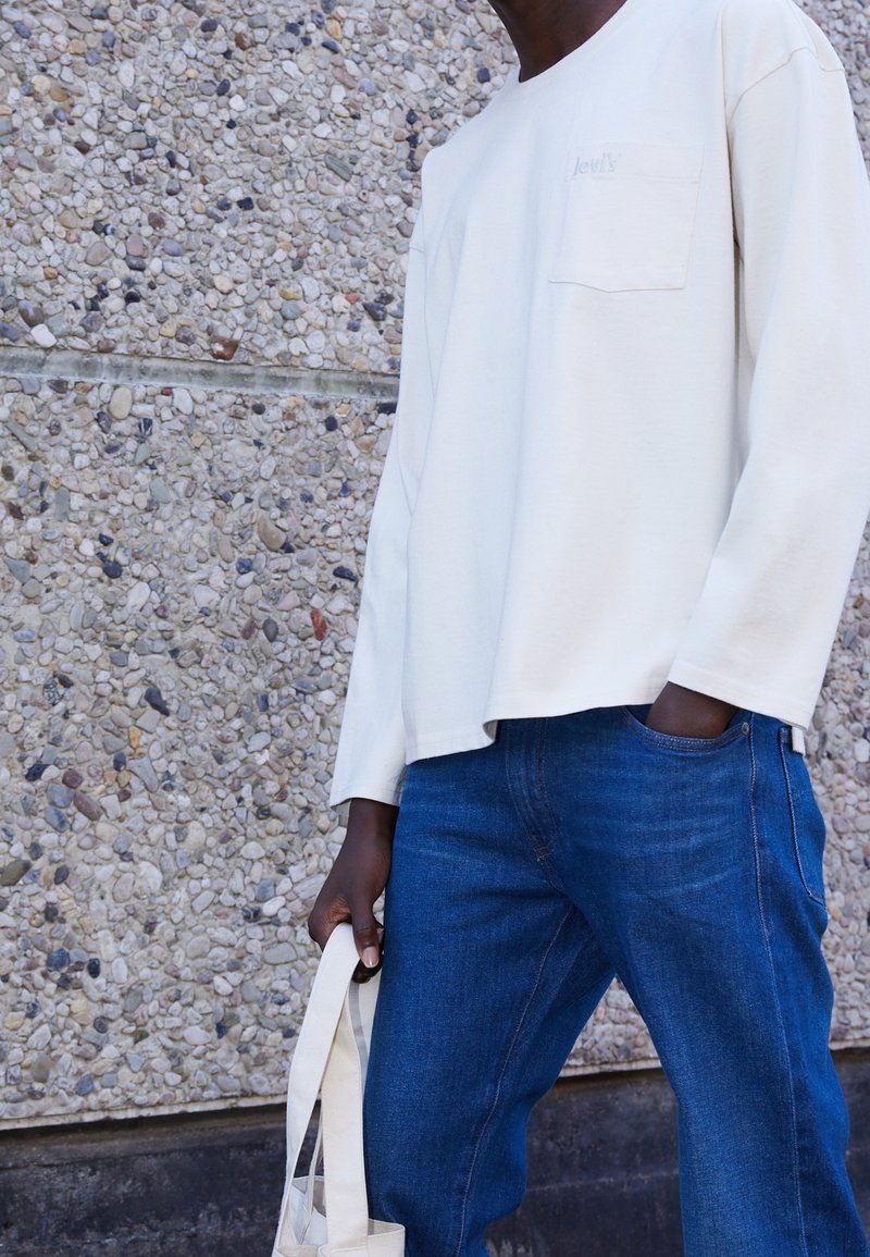 Chemise claire à manches longues avec une poche poitrine et un branding subtil, associée à un jean en denim bleu foncé et un sac tote beige.