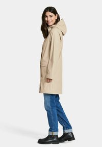 Beige vedenpitävä takki hupulla, jossa suora leikkaus, pitkät hihat ja etutaskut, yhdistetty sinisiin farkkuihin ja mustiin saappaisiin.