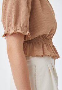 Blusa marrone con texture, maniche a sbuffo e orlo arricciato. Presenta una vita arricciata. Abbinata a pantaloni beige chiaro, che mostrano un profilo laterale.