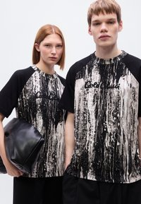 Μαύρα και λευκά γραφικά T-shirts με ραβδωτό σχέδιο, που διαθέτουν στρογγυλή λαιμόκοψη και κοντά μανίκια. Μοντέλο που κρατάει μια μαύρη τσάντα.