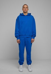 Modra bombažna kapuca s sprednjo žepom in vrvico, v kombinaciji z usklajenimi modrimi trenirka, ki imajo elastične manžete. Oblečeni so s belimi športniki.