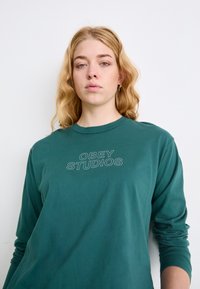 Dugo rukava, zeleni pamučni majica s laganim sivo-smešanim logotipom "OBEY STUDIOS" u blok fontu na prednjoj strani. Mekan, ležeran dizajn.