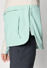 Lahka jakna ima spodnji del v zeleni meti, zgornji del v kremni barvi, stransko žep s potegom in ukrivljen rob. Material deluje gladko in utilitarno.