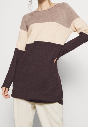 Kobieta ubrana w sweter z długim rękawem z kolorowymi pasami w poziomie w kolorach brązowym, beżowym i ciemnobrązowym, zestawiony z jasnymi spodniami.