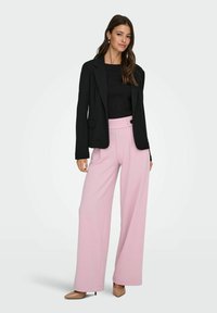 Black tailored blazer over a fitted black top, paired with high-waisted, wide-leg pastel pink trousers. Features a button detail. Neutral heels.