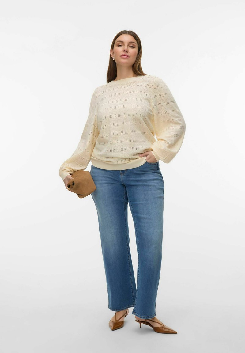 Pull en maille crème clair oversize avec motifs ouverts, associé à un jean droit bleu moyen. Tenant une pochette marron et portant des talons beige.