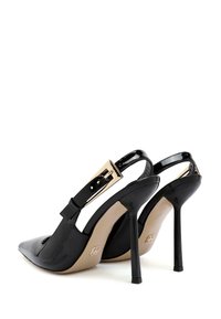 Zapatos de tacón alto de charol negro con abertura en la parte trasera, punta afilada y tira ajustable en el tobillo con herrajes en tono dorado.