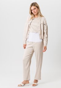Lichtbeige jas met knopen en zak, gelaagd over een witte top. Gecombineerd met bijpassende beige broek en witte instappers.