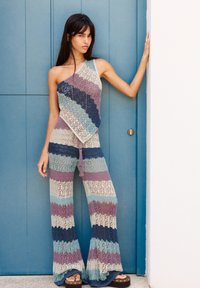Gehaakt twee-delig outfit in strepen van lichtblauw, paars en crème, met een één-schouder top en uitlopende broek met franjes.
