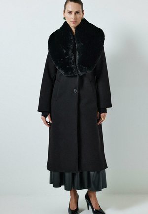 Moteris vilkinti ilgą juodą paltą su dideliu dirbtinio kailio apykakle, juodą sijoną, juodus aukštakulnius batus, ir auksines auskarus bei žiedus, stovinti prieš paprastą foną.