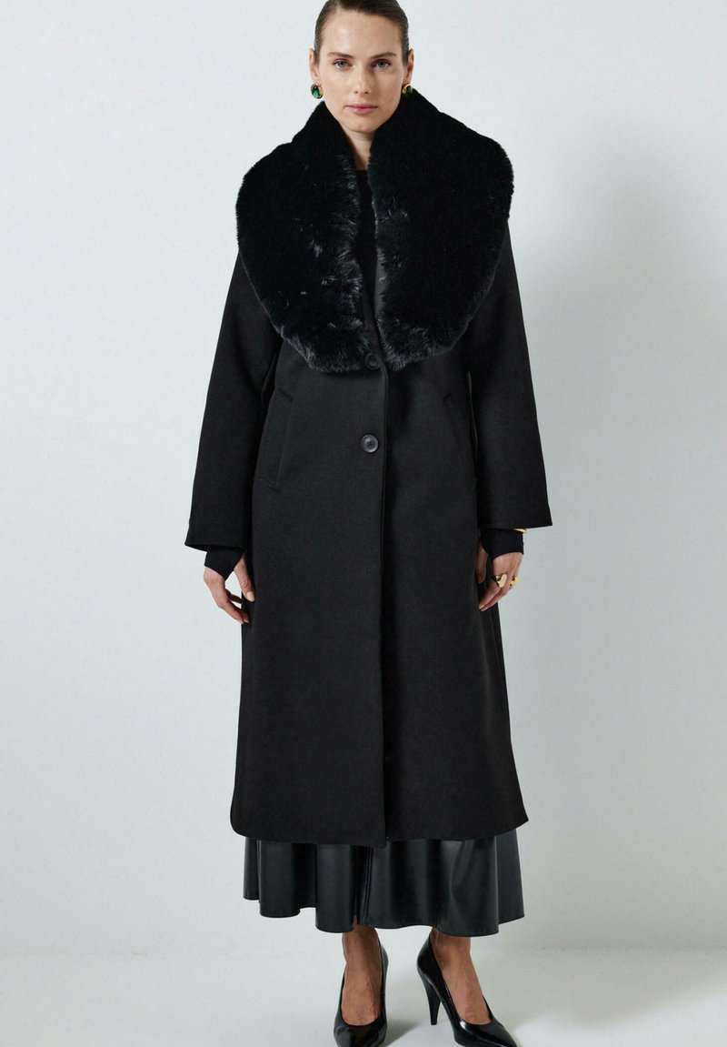 Vrouw in een lange zwarte jas met een grote nep bontkraag, zwarte rok, zwarte hoge hakken, en gouden oorbellen en ringen, staande tegen een effen achtergrond.