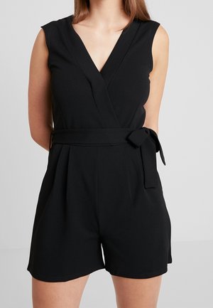 Femme portant une combinaison noire sans manches à col en V avec une ceinture nouée à la taille, sur un fond gris uni.
