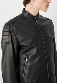 Black leather jacket with a stand collar, zip closure, and padded shoulder accents. Features an exterior pocket with a silver zipper.