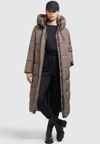 Long padded coat in brown with a hood, featuring a two-way zipper and logo details. Worn over a black outfit and paired with black boots.