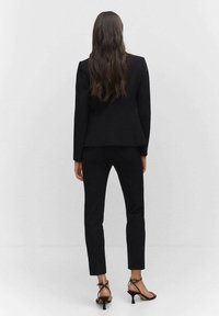 Blazer negro a medida sobre pantalones a juego, ambos fabricados con tejido texturizado. Tacones negros con tiras complementan el conjunto. Diseño simple y elegante.