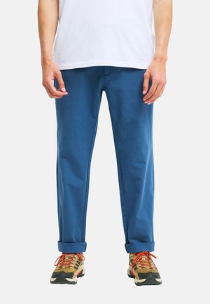 Homme portant un pantalon bleu retroussé, un T-shirt blanc et des chaussures de randonnée beiges avec des lacets orange, debout devant un fond uni.