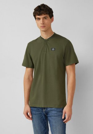 Jonge man staand, gekleed in een olijfgroene henley met korte mouwen en een klein borstlogo, blauwe spijkerbroek, neutrale grijze achtergrond.