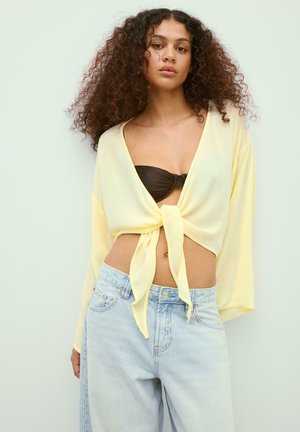 Žena s kovrčavom kosom nosi svijetložuti top dugih rukava s čvorom sprijeda preko crnog bralett-te i širokih svijetloplavih traperica, stoji na običnoj pozadini.