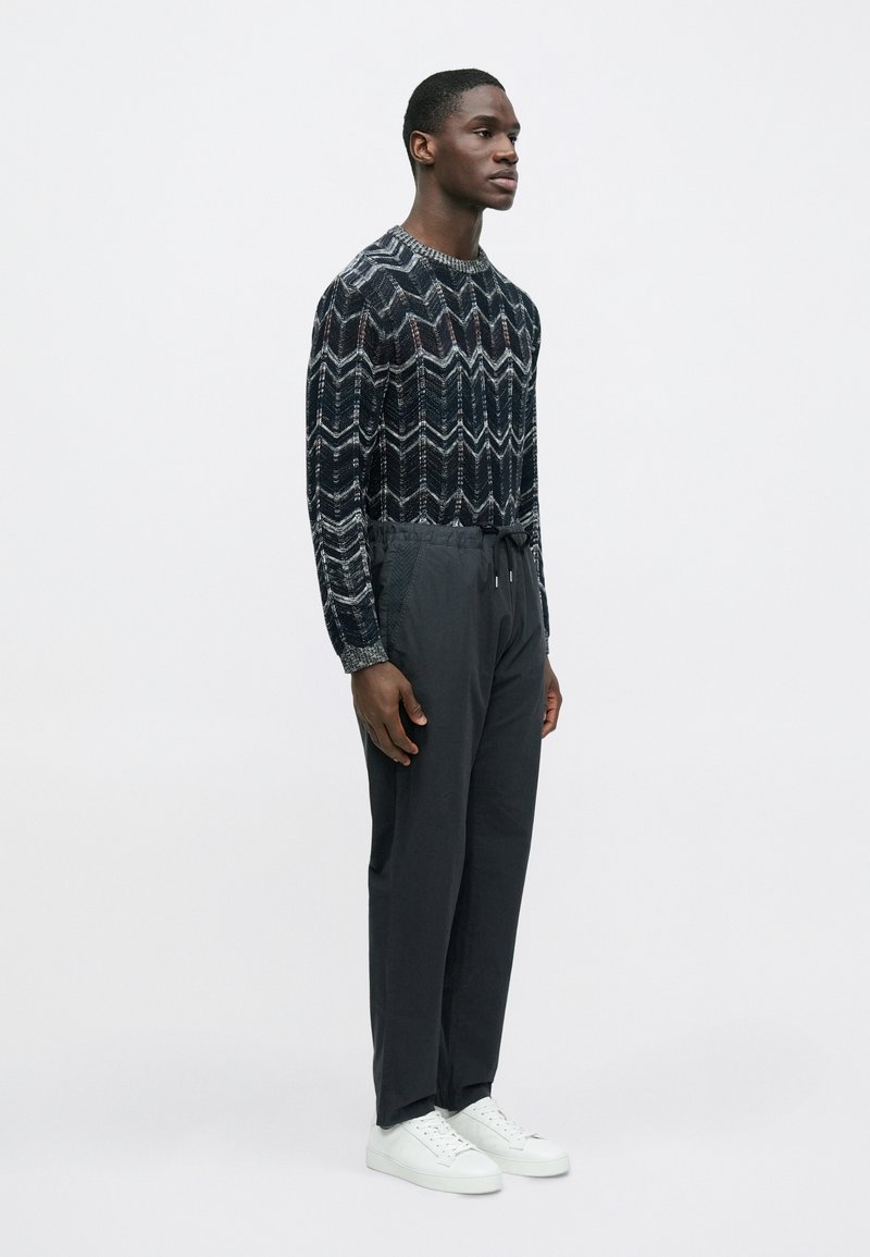 Pull en maille à motifs noirs et blancs avec un design en zigzag, associé à un pantalon gris foncé et des baskets blanches.
