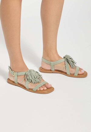 Feet wearing flat, open-toe sandals with light green straps and decorative tassels on a light brown sole against a plain background.