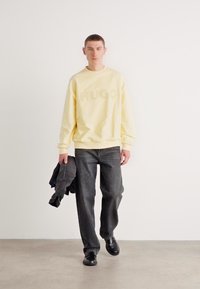 Sweatshirt amarelo claro com o logótipo "HUGO", jeans escuros e sapatos pretos; modelo segura um casaco escuro. Design simples, corte descontraído, textura suave.