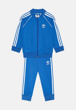 Blauwe Adidas trainingsoutfit met witte drie-streepjes op de mouwen en broek. Beschikt over een jack met ritssluiting en geribbelde manchetten. Gemaakt van soepel materiaal.