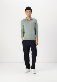 Light green zip-neck sweater with ribbed cuffs and hem, paired with navy trousers and white sneakers. Simple, casual outfit on a model.