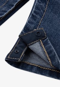 Un tissu en denim bleu foncé présente un pli avec une texture intérieure claire contrastante. Détails de couture visibles et accents en métal circulaire.