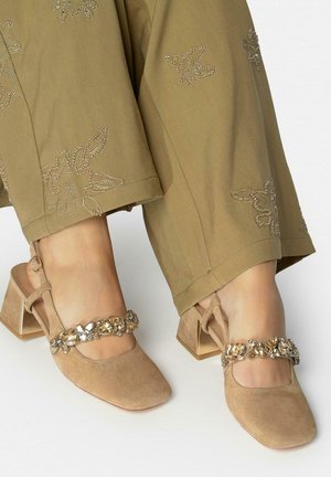 Tacones de bloque de ante beige con tiras decorativas con joyas, usados con pantalones caqui bordados.