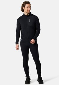 Black zip-up athletic jacket with long sleeves, high collar, and side zip pockets, paired with black fitted leggings and sneakers.