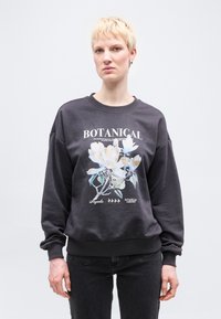 Άτομο με κοντά ξανθά μαλλιά που φοράει ένα σκούρο φούτερ με μεγάλο λουλουδένιο σχέδιο και κείμενο "BOTANICAL" στέκεται μπροστά σε ένα απλό φόντο.