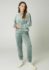 Set di abbigliamento loungewear in velluto verde chiaro. Felpa con cappuccio e cordino, maniche lunghe. Pantaloni con polsini elasticizzati. Sneakers bianche con dettagli testurizzati.