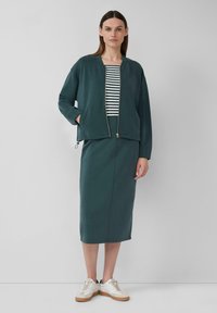 Veste zippée vert foncé superposée à un haut rayé blanc et marine, associée à une jupe assortie et des baskets beiges. Texture de tissu lisse.